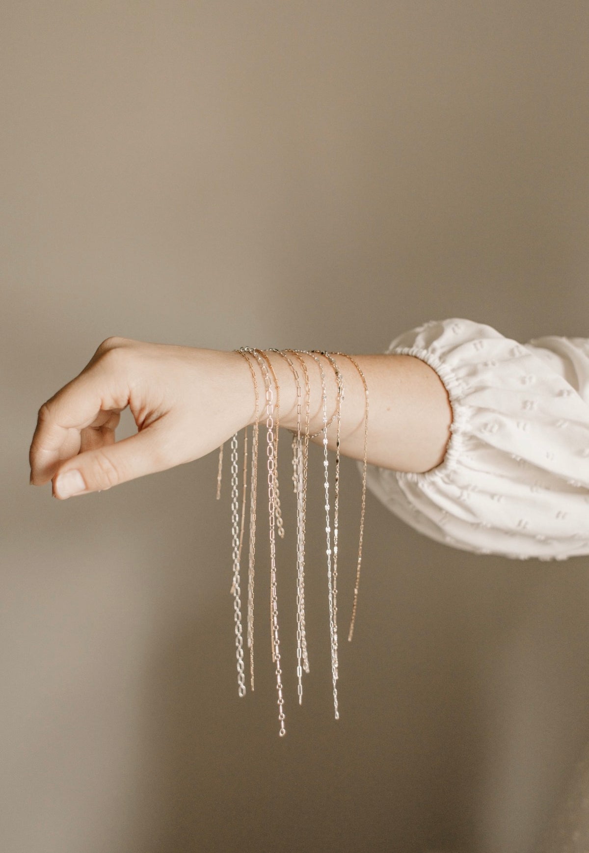 A close-up of a person's wrist and hand wearing multiple delicate rose gold chain bracelets with long dangling fringe, paired with a white long-sleeve blouse against a neutral beige background.