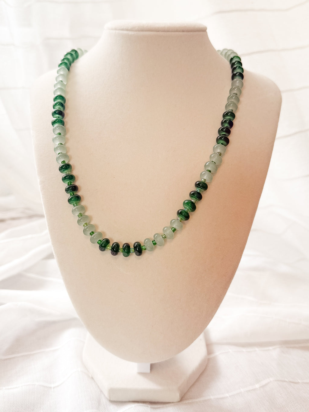A beaded necklace made of alternating dark green and light mint green beads, displayed on a cream-colored jewelry bust against a draped white fabric background.