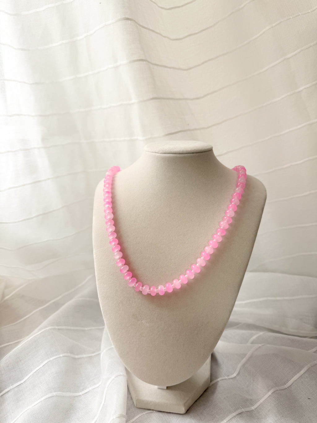 A pink beaded necklace displayed on a cream-colored jewelry bust against a white textured fabric backdrop.